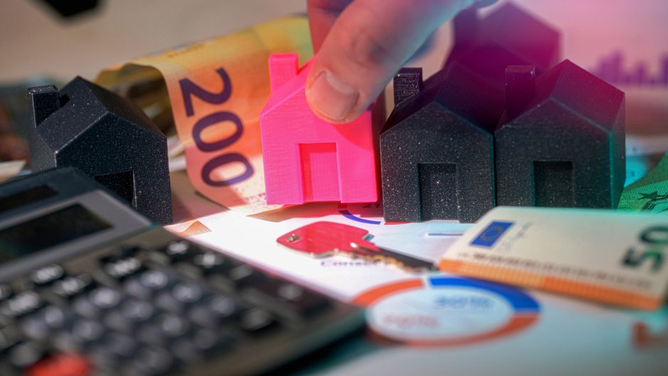 A close-up view of a person's finger placing a small, bright pink plastic house model among larger black miniature house models on a cluttered surface. The scene includes various Euro banknotes, such 